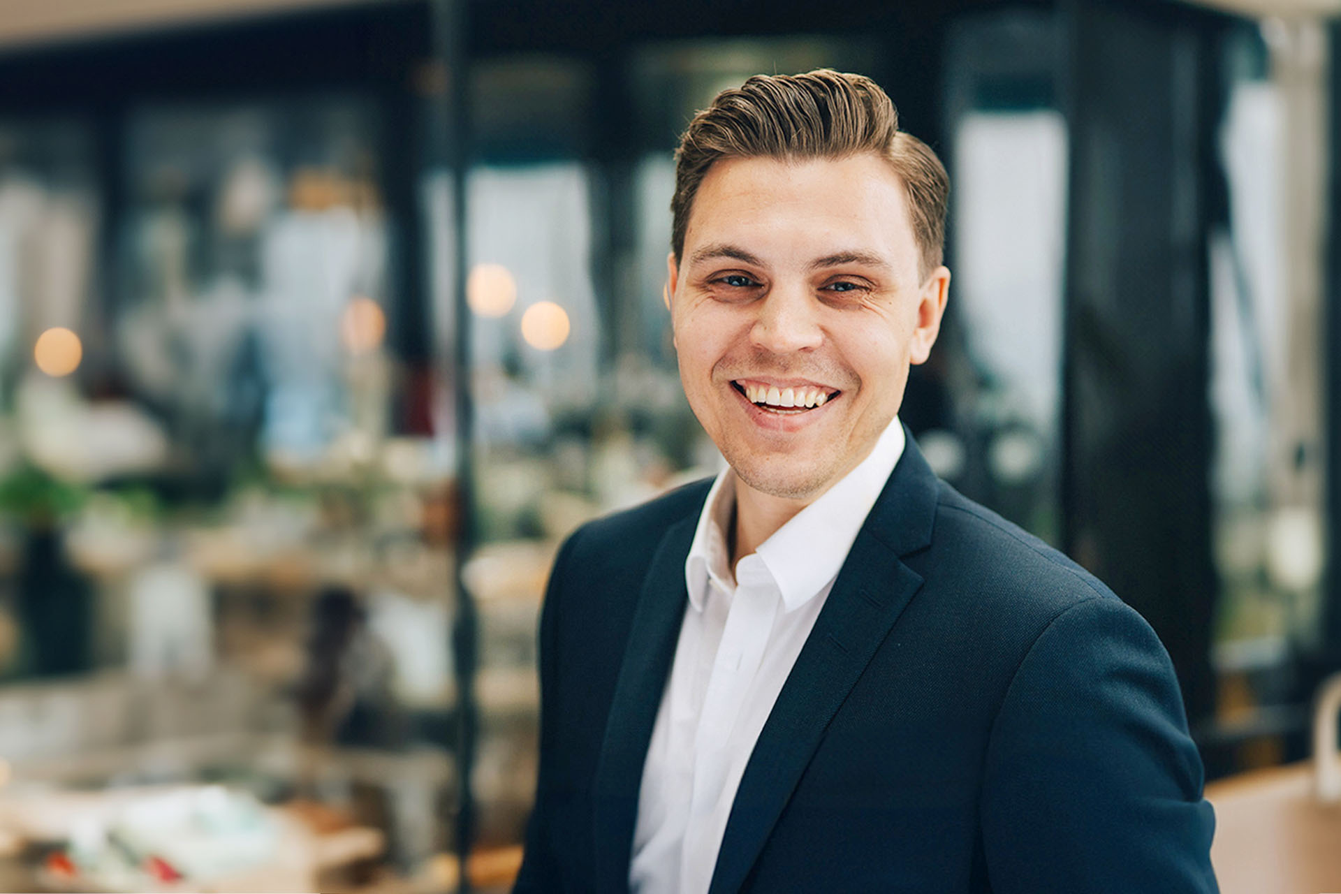 En ung man i en mörk kostym och vit skjorta ler mot kameran i en ljus och modern miljö.